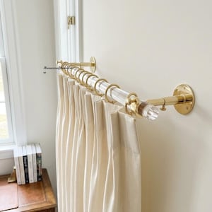 May include: A clear acrylic curtain rod with polished brass wall mounts and matching brass rings. The rod holds pleated off-white curtains. The text Homexcrafthardware is visible on the wall near the left mounting bracket.