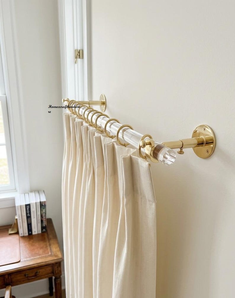 May include: A clear acrylic curtain rod with polished brass wall mounts and matching brass rings. The rod holds pleated off-white curtains. The text Homexcrafthardware is visible on the wall near the left mounting bracket.