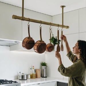 May include: A ceiling-mounted brass pot rack featuring a horizontal bar suspended by two vertical rods. Three copper saucepans with long handles and two wooden kitchen utensils hang from the rack using S-shaped metal hooks in a kitchen.