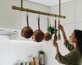 May include: A ceiling-mounted brass pot rack featuring a horizontal bar suspended by two vertical supports. Three copper saucepans and two wooden kitchen utensils hang from the bar using S-shaped hooks in a modern kitchen setting.