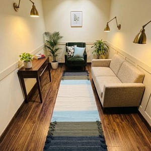 Puede incluir: Una alfombra larga y rectangular con un patrón de rayas en tonos azules, blancos y verdes. La alfombra está colocada sobre un suelo de madera en una habitación con un sofá, una silla y un escritorio. La habitación está decorada con plantas y apliques de pared.