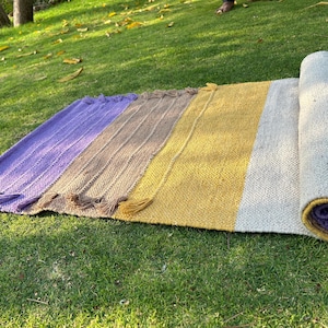 Puede incluir: Una alfombra enrollada multicolor con un diseño a rayas. La alfombra presenta secciones de color púrpura, marrón, amarillo y blanco. La parte enrollada revela un interior púrpura con un borde dorado. Las borlas adornan el borde de la alfombra.