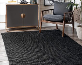 May include: A large, rectangular black rug with a textured, ribbed surface. The rug is made of a woven material and is placed on a hardwood floor. The rug is in a living room setting, near a wooden chair and cabinet.