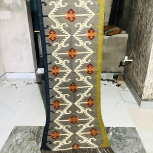 May include: A long, rectangular runner rug featuring a grey base with a repeating geometric pattern in white and orange. The rug has a dark blue border on one side and a mustard yellow border on the other, finished with short, dark brown fringe.