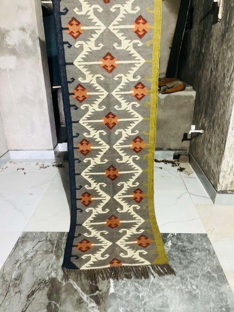May include: A long, rectangular runner rug featuring a grey base with a repeating geometric pattern in white and orange. The rug has a dark blue border on one side and a mustard yellow border on the other, finished with short, dark brown fringe.