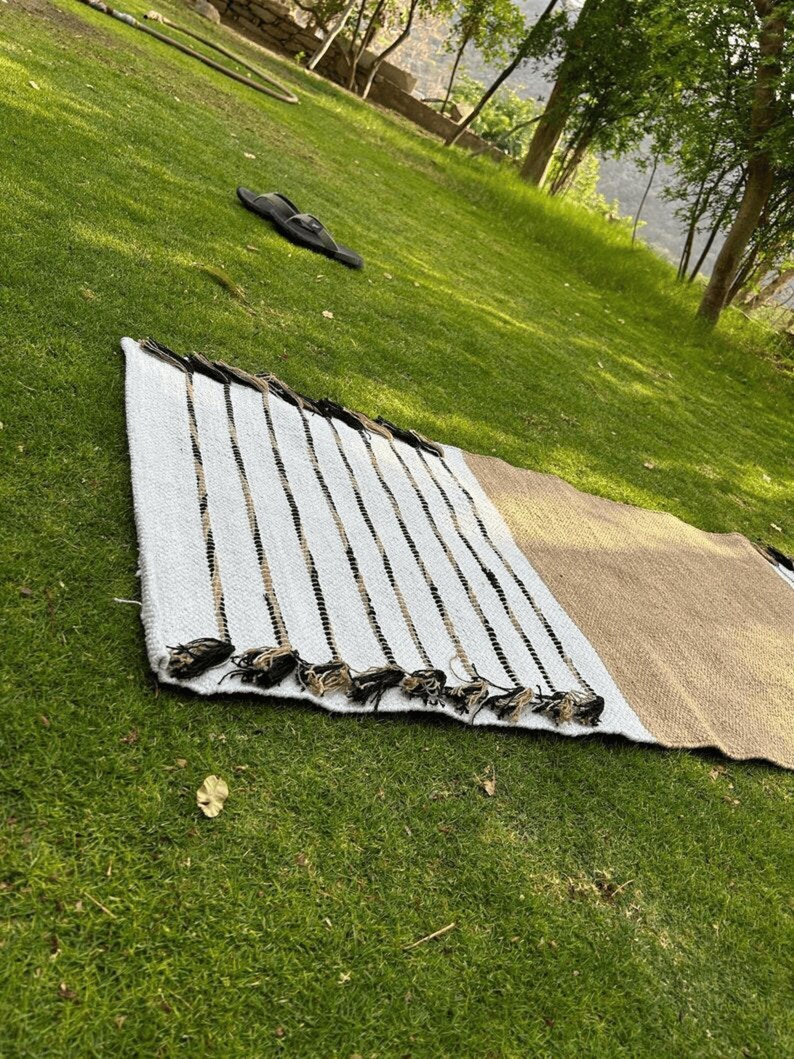 Puede incluir: Una alfombra rectangular tejida con un patr&oacute;n de rayas en blanco, beige y negro, con borlas negras a lo largo de un borde. La alfombra est&aacute; sobre hierba verde, con un par de sandalias negras en el fondo.