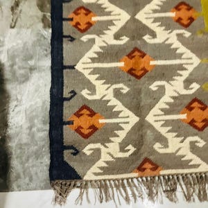 May include: A rectangular woven rug featuring a grey background with a repeating geometric pattern of white zig-zag lines and orange diamond shapes. The rug has a dark navy blue border along the left side and a fringe of light brown tassels at the bottom.