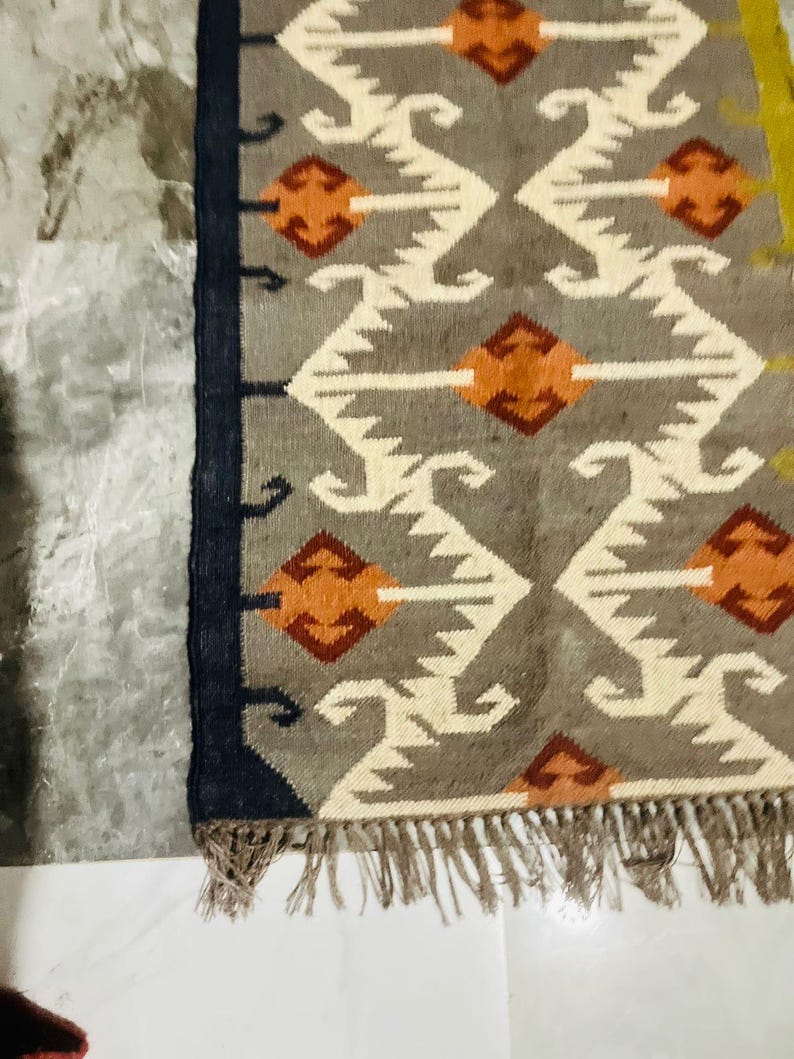 May include: A rectangular woven rug featuring a grey background with a repeating geometric pattern of white zig-zag lines and orange diamond shapes. The rug has a dark navy blue border along the left side and a fringe of light brown tassels at the bottom.