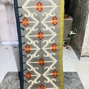 May include: A long, rectangular runner rug featuring a grey background with a repeating geometric pattern in white, orange, and dark red. The rug has a dark blue border on the left side and a mustard yellow border on the right side.