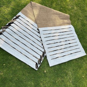 Puede incluir: Una alfombra rectangular con una secci&oacute;n superior marr&oacute;n y una secci&oacute;n inferior blanca con rayas negras. La alfombra tiene borlas en un extremo y se exhibe sobre hierba verde.