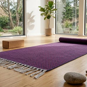 Puede incluir: Una esterilla de yoga rectangular de color morado con un patrón de diamantes texturizado y flecos blancos en un extremo. Un bloque de yoga rectangular de corcho marrón se sitúa a la izquierda. La esterilla está parcialmente enrollada sobre un suelo de madera clara.