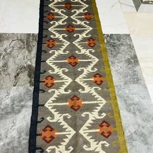 May include: A long rectangular runner rug featuring a grey background with a repeating geometric pattern in white and burnt orange. The rug has a solid black border on one side, a solid olive green border on the other, and fringed ends in grey.