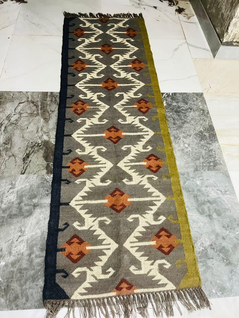 May include: A long rectangular runner rug featuring a grey background with a repeating geometric pattern in white and burnt orange. The rug has a solid black border on one side, a solid olive green border on the other, and fringed ends in grey.