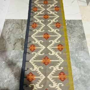 May include: A rectangular runner rug featuring a grey base with a repeating geometric pattern in white and orange. The rug has a dark blue border on the left side, a mustard yellow border on the right side, and fringed ends at the top and bottom.