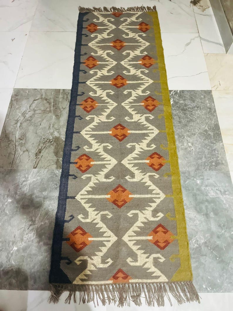 May include: A rectangular runner rug featuring a grey base with a repeating geometric pattern in white and orange. The rug has a dark blue border on the left side, a mustard yellow border on the right side, and fringed ends at the top and bottom.