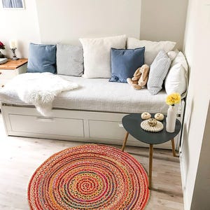 Puede incluir: Un espacio de vida acogedor con un sof&aacute; cama blanco, cojines azules y blancos y una alfombra redonda multicolor. La alfombra tiene un dise&ntilde;o en espiral con tonos rojos, naranjas y amarillos. Una peque&ntilde;a mesa auxiliar con un jarr&oacute;n y flores amarillas.