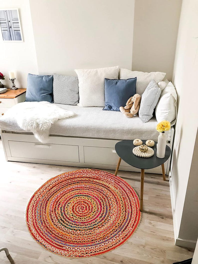 Puede incluir: Un espacio de vida acogedor con un sof&aacute; cama blanco, cojines azules y blancos y una alfombra redonda multicolor. La alfombra tiene un dise&ntilde;o en espiral con tonos rojos, naranjas y amarillos. Una peque&ntilde;a mesa auxiliar con un jarr&oacute;n y flores amarillas.