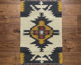 May include: Rectangular woven rug with a geometric pattern in cream, gray, yellow, and red. The rug has a fringed edge and is displayed on a wooden floor. The design features symmetrical shapes and a central medallion.