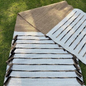 Puede incluir: Dos alfombras tejidas con una paleta de colores neutros. Una alfombra es principalmente blanca con rayas horizontales negras y beige y flecos. La otra alfombra es beige con un borde blanco. Las alfombras se exhiben sobre hierba verde.