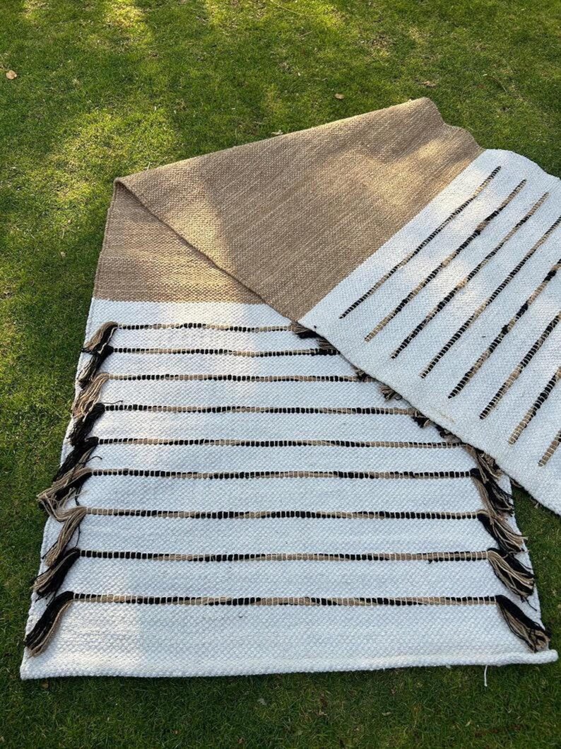 Puede incluir: Dos alfombras tejidas con una paleta de colores neutros. Una alfombra es principalmente blanca con rayas horizontales negras y beige y flecos. La otra alfombra es beige con un borde blanco. Las alfombras se exhiben sobre hierba verde.