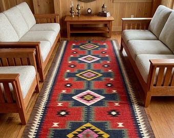 May include: A long, rectangular red rug with a geometric pattern in navy blue, yellow, pink, and white. The rug has a brown border and fringe. The rug is placed between two light-colored sofas and wooden furniture.