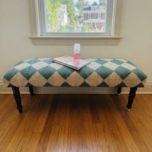 May include: A rectangular bench featuring a teal and cream diamond pattern upholstery, supported by four dark brown turned wooden legs. A spiral-bound notebook and a small, clear plastic bottle with a pink liquid are placed on the bench seat.