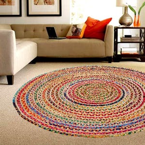 Puede incluir: Una alfombra redonda trenzada de colores vibrantes, con c&iacute;rculos conc&eacute;ntricos de rojo, azul, verde y amarillo. La alfombra est&aacute; colocada sobre una moqueta de tono neutro, cerca de un sof&aacute; beige con cojines naranjas. El di&aacute;metro es de unos 150 cm.