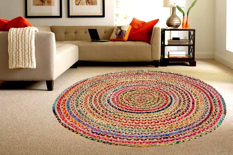 Puede incluir: Una alfombra redonda trenzada de colores vibrantes, con c&iacute;rculos conc&eacute;ntricos de rojo, azul, verde y amarillo. La alfombra est&aacute; colocada sobre una moqueta de tono neutro, cerca de un sof&aacute; beige con cojines naranjas. El di&aacute;metro es de unos 150 cm.