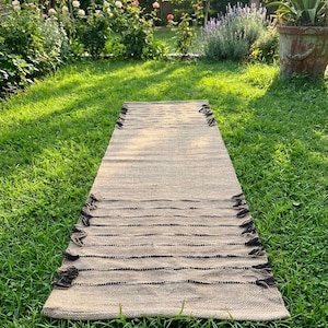 Puede incluir: Una alfombra rectangular de yute beige con rayas horizontales negras y flecos en los extremos. La alfombra está colocada sobre hierba verde, con un jardín y árboles al fondo. Un elemento de decoración para el hogar de tonos neutros y naturales.