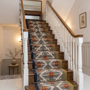 May include: A staircase runner featuring a grey, olive green, and orange geometric pattern with a fringed edge. The runner is secured to wooden steps with metallic rods, framed by white balusters and a wooden handrail leading to a landing.