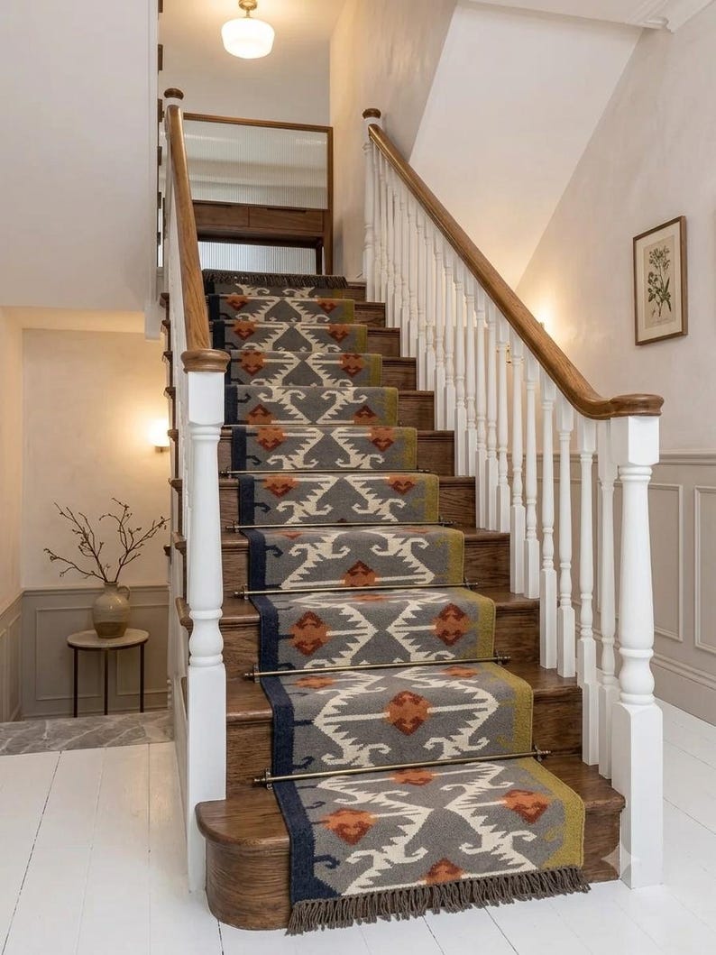 May include: A staircase runner featuring a grey, olive green, and orange geometric pattern with a fringed edge. The runner is secured to wooden steps with metallic rods, framed by white balusters and a wooden handrail leading to a landing.