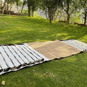 Puede incluir: Una alfombra rectangular para exteriores con un centro de fibra natural y extremos a rayas blancas con detalles en negro. La alfombra est&aacute; colocada sobre una hierba verde, lo que sugiere un picnic o un ambiente de relajaci&oacute;n al aire libre.