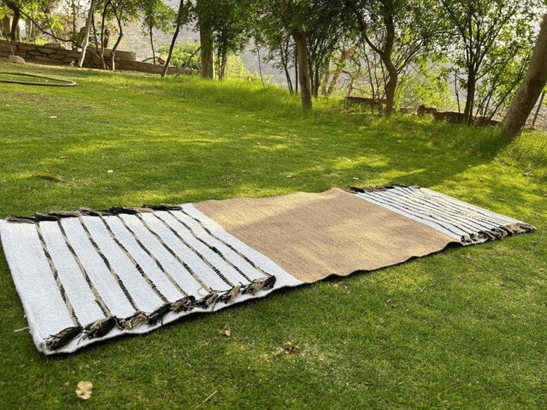 Puede incluir: Una alfombra rectangular para exteriores con un centro de fibra natural y extremos a rayas blancas con detalles en negro. La alfombra est&aacute; colocada sobre una hierba verde, lo que sugiere un picnic o un ambiente de relajaci&oacute;n al aire libre.