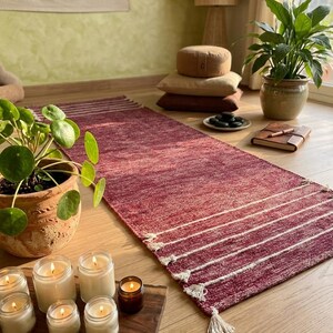 Puede incluir: Una alfombra rectangular tejida de color rojo intenso con rayas horizontales blancas y borlas decorativas en ambos extremos. La alfombra tiene una textura tejida visible y está colocada sobre un suelo de madera clara.