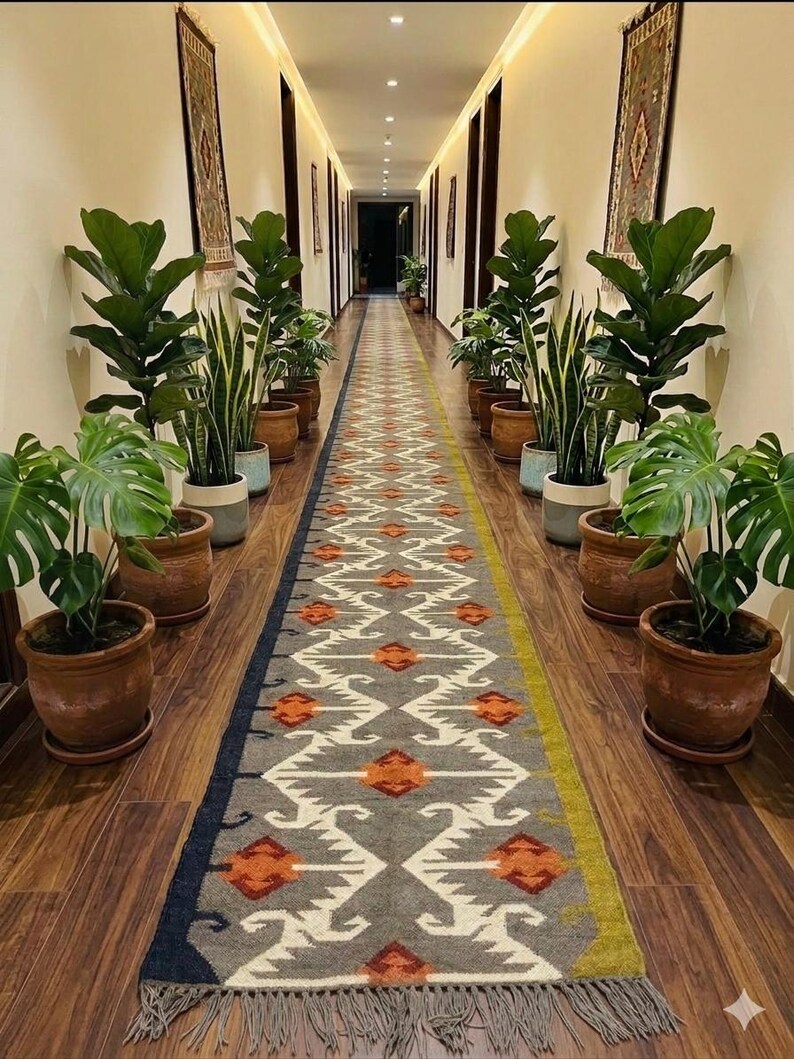 May include: A long, rectangular runner rug featuring a grey base with a repeating geometric pattern in white, orange, and dark blue. The rug has a fringed edge and is positioned in a hallway lined with various potted green plants on both sides.