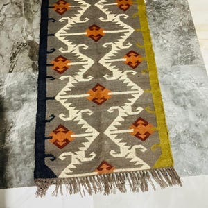May include: A rectangular runner rug featuring a grey base with a repeating white geometric pattern and orange diamond-shaped accents. The rug has a solid black border on one side, a solid olive green border on the other, and frayed fringe ends.