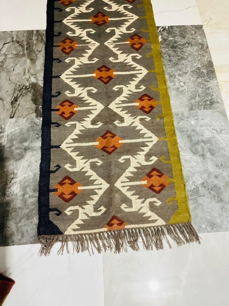 May include: A rectangular runner rug featuring a grey base with a repeating white geometric pattern and orange diamond-shaped accents. The rug has a solid black border on one side, a solid olive green border on the other, and frayed fringe ends.