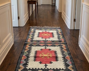 Puede incluir: Una alfombra rectangular con un patrón geométrico en un pasillo. La alfombra presenta un cuadrado rojo central con un diseño amarillo, rodeado por un borde blanco y un borde negro y gris. La alfombra tiene flecos en ambos extremos.