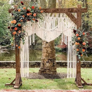 Puede incluir: Un arco de boda de madera decorado con macramé color crema y arreglos florales de flores naranjas y burdeos. El arco está instalado al aire libre sobre una zona de césped, con un lago al fondo.