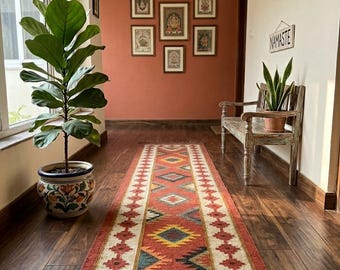 May include: A hallway featuring a long, rectangular rug with a geometric pattern in red, yellow, and black. The rug is on a dark wood floor. A potted plant and framed artwork adorn the walls, creating a warm, inviting atmosphere.