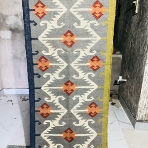 May include: A rectangular woven rug featuring a grey background with a repeating geometric pattern in white, orange, and dark red. The rug has a dark blue border on the left side and a mustard yellow border on the right side, with fringe at the top.