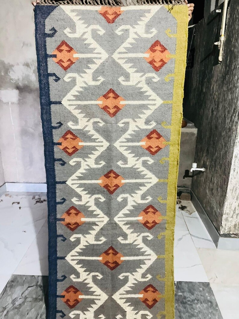 May include: A rectangular woven rug featuring a grey background with a repeating geometric pattern in white, orange, and dark red. The rug has a dark blue border on the left side and a mustard yellow border on the right side, with fringe at the top.