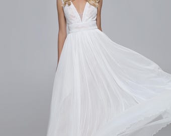 May include: A white wedding dress with a V-neckline and flowing skirt. The dress features delicate lace detailing on the bodice and a floral crown. The model is smiling, with blonde hair.