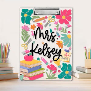 May include: A clear clipboard with a metal clip, decorated with colourful flowers, pencils, scissors, and books. The name "Mrs. Kelsey" is written in black script. A+ grade is shown on a yellow paper.