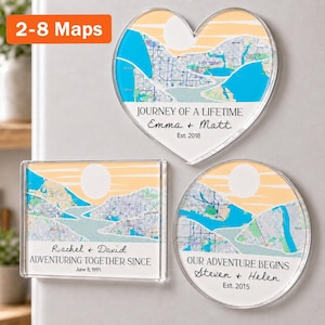 May include: Three clear acrylic magnets in heart, rectangular, and circular shapes featuring stylized maps with blue water and peach skies. Text includes JOURNEY OF A LIFETIME Emma + Matt Est. 2018, Rachel + David ADVENTURING TOGETHER SINCE June 8, 1991, and OUR ADVENTURE BEGINS Steven + Helen Est. 2015.
