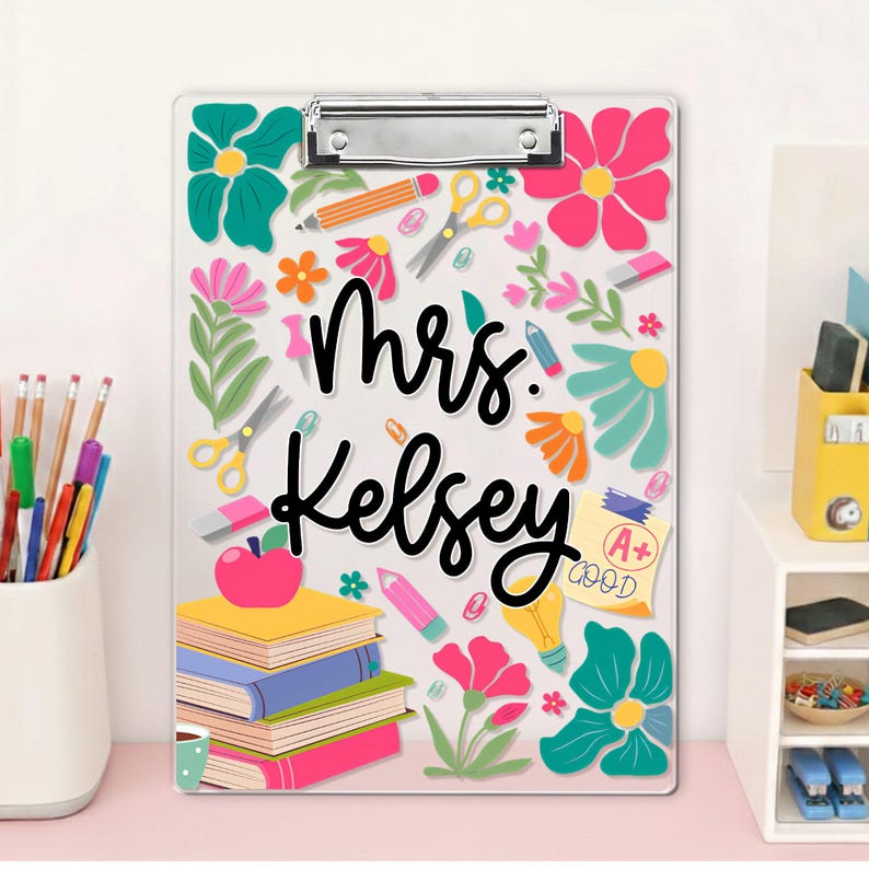 May include: A clear clipboard decorated with colourful flowers, pencils, scissors, and books. The name "Mrs. Kelsey" is written in black script. A stack of books with a red apple on top is also featured.