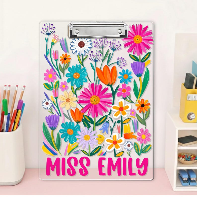 May include: A clear clipboard adorned with a colourful floral pattern, including pink, orange, blue, and yellow blooms. The words "MISS EMILY" are printed in pink at the base. A silver clip secures papers at the top.