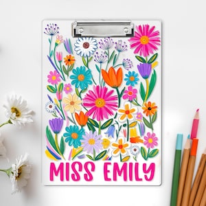 May include: A white clipboard adorned with a colourful floral pattern, including pink, orange, blue, and yellow flowers. The text "MISS EMILY" is in pink. A silver clip secures the paper. Pencils are in the background.