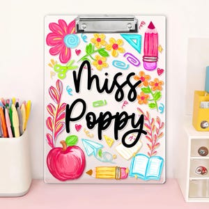 May include: A clear clipboard with a metal clip, decorated with colourful school-themed illustrations. The design includes flowers, an apple, a pencil, and the name "Miss Poppy" in black script. The clipboard is a standard A4 size.