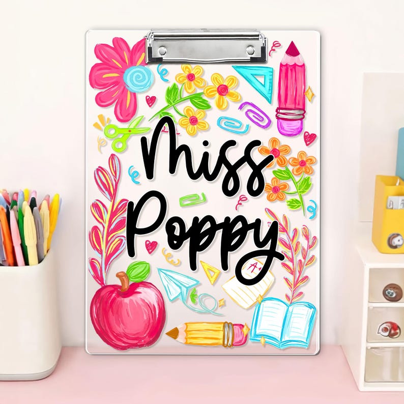 May include: A clear clipboard with a metal clip, decorated with colourful school-themed illustrations. The design includes flowers, an apple, a pencil, and the name "Miss Poppy" in black script. The clipboard is a standard A4 size.